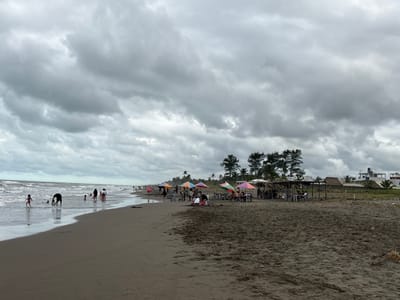 A cloudy beach scene shows a wide stretch of dark sand meeting the ocean. Several people are wading and playing in the water. Further down the beach, groups gather under colorful umbrellas and thatched structures. The sky is overcast with dense, grey clouds, suggesting a potentially stormy day. Beyond the beach area, some vegetation and a few buildings are visible.