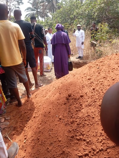 The image depicts an outdoor burial site in Okpatu-Ukehe, Nigeria, under bright daylight conditions. In the foreground, a large mound of reddish-brown, freshly excavated soil occupies the right half of the frame.

Approximately twelve individuals are visible, primarily gathered around a white casket with gold-colored handles situated on the ground in the mid-left. A person wearing a long, flowing purple robe, a matching head covering, and a cape-like garment, seen from the back, stands near the center, holding a black handbag in their left hand. This individual is positioned adjacent to the excavated grave pit, which is partially visible behind the soil mound.

To the left of the casket, several men are partially visible from the back and sides. One wears a yellow t-shirt and grey shorts; another wears a black t-shirt and dark shorts, holding a dark object, possibly a mobile phone. Another man wears a patterned black t-shirt and red trousers. Their lower legs and feet are visible, some in sandals, others appearing barefoot.

To the right of the person in purple, a man in a white tunic-style top, white trousers, and a dark cap, with a beard, stands near the edge of the grave pit, holding a mobile phone in his right hand as if recording or photographing the scene. Further back and to his right, another man in a dark patterned tank top observes the scene. Several other individuals, some in white attire, are visible in the background between the casket and the recording man.

The background features sparse vegetation, including trees and shrubs, indicating an undeveloped or semi-rural setting. Shadows are distinct, suggesting a high sun angle. The scene collectively represents a funeral or burial ceremony in progress.