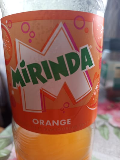 This is a close-up, eye-level, vertical shot of a plastic bottle of Mirinda Orange soda. The bottle is filled with orange liquid and has a prominent orange and white label with green lettering that spells out "MIRINDA" in large font. The label also features circular orange and pink designs, as well as stylized images of orange slices and splashes. Below the "MIRINDA" text, the word "ORANGE" is clearly visible in white capital letters, followed by smaller text that reads "LICIOUS & FLAVOURFUL TASTE". The bottle is positioned on a surface covered with a pink and green floral patterned cloth. The background is blurred, showing hints of green and dark colors, suggesting an indoor setting. The image appears to be taken in Zīlāni, Latvia, given the context of the prompt.