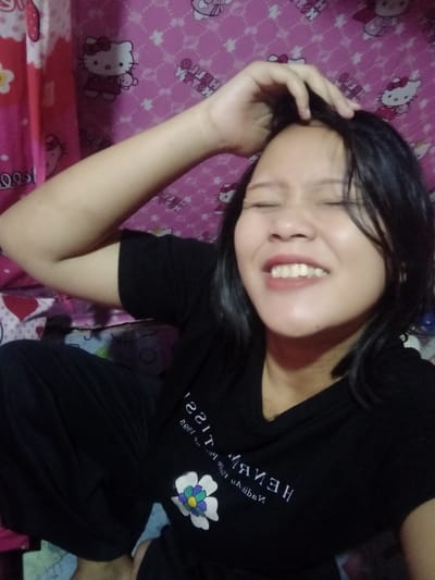 A woman poses for a selfie with a hand on her head. She wears a black t-shirt with a white flower design and script. Her dark hair is damp. The background features a pink and white Hello Kitty patterned wall, and a pink curtain can be seen to the left. She has a slight smile and her eyes are closed, creating a squinting effect. She appears to be in a relaxed setting, possibly at home.