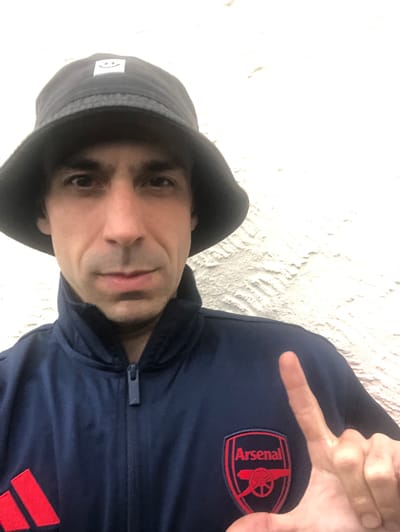 An indoor portrait depicts a male individual located in Shotton, United Kingdom. The subject, with dark hair and a fair complexion, faces the camera directly. He wears a dark bucket hat featuring a white square patch with a black smiley face emblem on the front. His attire consists of a dark blue, zippered track jacket, which displays an embroidered red and blue Arsenal Football Club crest on the left chest and a partial red Adidas three-stripe logo on the lower left. The subject's right hand is raised, forming an 'L' shape with an extended index finger and horizontally outstretched thumb. The background is a uniformly illuminated, textured white wall.
