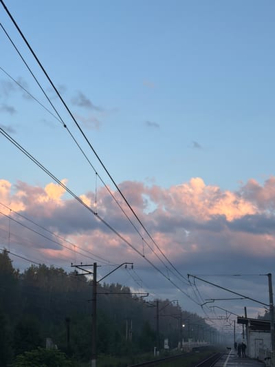 The image depicts a view of a train station platform and tracks in Orekhovo, Russia, during twilight. The sky is a pale blue, with wispy clouds tinged pink and grey, suggesting either sunrise or sunset. Overhead, a complex network of power lines for an electric train is visible, supported by utility poles. The platform itself is partially visible on the right, with a building structure and a few silhouetted figures standing on it, hinting at the presence of passengers or station staff. The tracks stretch into the distance, bordered by lush green foliage and trees, creating a natural backdrop to the industrial infrastructure. The overall mood is serene and quiet, typical of the transitional light of dawn or dusk.