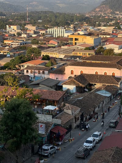 The image is an aerial view of a section of La Esperanza, Honduras, characterized by a mix of residential and commercial buildings with clay tile roofs. The buildings are of varying heights and colors, including yellow, pink, and white, suggesting a historical urban center. A street runs diagonally across the middle ground, featuring parked vehicles, including several white SUVs, and motorcycles. Pedestrians are visible on the sidewalks and near vehicles, indicating urban activity. A large Pepsi advertisement is visible on the side of one of the buildings. In the background, a forested hillside rises, partially obscured by a haze or mist. A construction crane is also visible in the background. The lighting suggests either early morning or late afternoon.