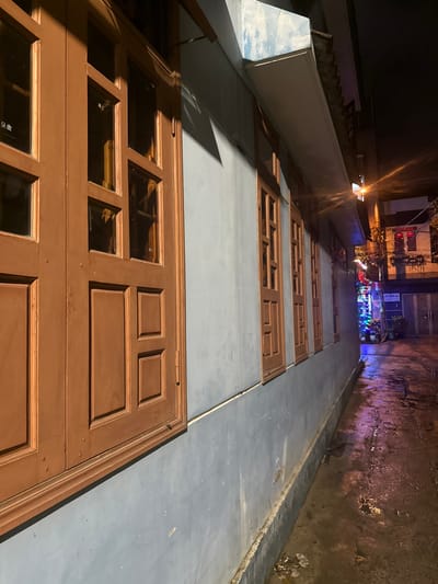 The image displays a night scene within Ngũ Hành Sơn Ward, Vietnam, focusing on the exterior of a building adjacent to a narrow, wet alleyway. The primary structure on the left features a light blue wall with a series of at least five closed, vertically oriented wooden windows, each having multiple brown frames and glass panes. A light-colored concrete overhang extends above the windows, casting a shadow onto the upper portion of the wall. The ground surface, appearing to be concrete or asphalt, is visibly wet, reflecting ambient light. A darker grey concrete base forms the lower section of the building wall. In the mid-ground, a street light emits an orange glow, creating lens flares and illuminating the wet ground. Further down the alley on the right, a darker building is visible with red lights, possibly internal illumination or signage. A brightly lit decorative display, featuring blue and white lights, is visible in the far background, positioned near several parked motorcycles or scooters. No individuals are identifiable within the scene.