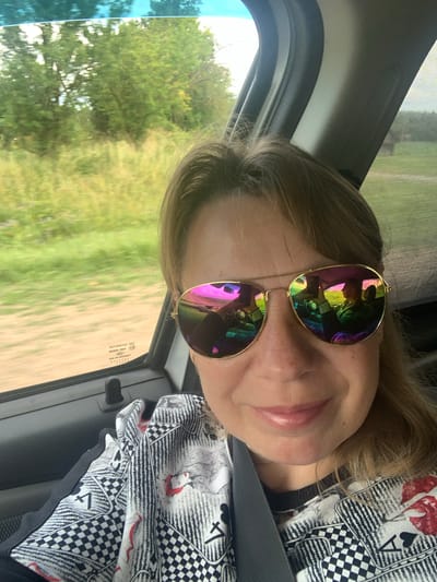 A woman is taking a selfie in a car. She is wearing aviator sunglasses with pink and green lenses. Her hair is light brown and she has on a black and white shirt with playing card motifs. The background shows greenery and trees, suggesting a rural setting. In the reflection of her sunglasses, another person is visible driving the car and holding a phone. The image appears to be taken during the daytime. The car's interior, specifically the door panel and window, are also visible.