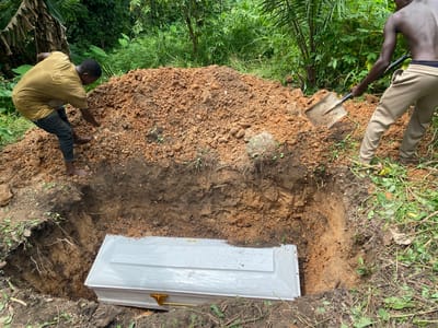 Two men are engaged in the process of burying a coffin in a grave dug in a rural area with dense vegetation. The grave is a rectangular pit in the earth, with a white coffin, adorned with gold-colored handles, already placed at the bottom. A pile of freshly dug reddish-brown soil is situated beside the grave. One man, wearing a yellow t-shirt and dark pants, is bending over the pile of soil, using a shovel to move it. The other man, shirtless and wearing light-colored sweatpants, is standing near the edge of the grave and also holding a shovel, seemingly in the act of excavating or leveling the soil. The surrounding environment is lush and green, with various trees and plants, suggesting an outdoor, likely remote, location in Igbara, Nigeria. The overall scene depicts a burial ritual taking place in a natural, uncultivated setting.