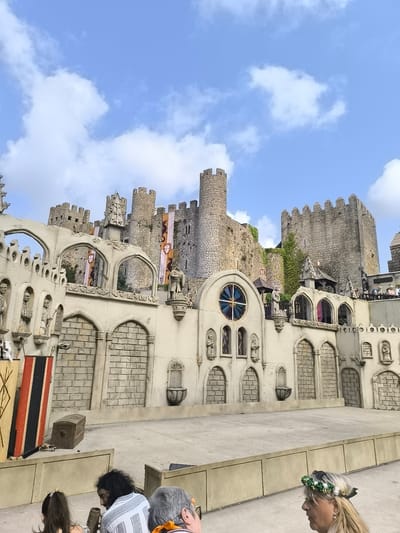 The image depicts a re-enactment of a medieval scene outside a castle, likely at the Óbidos Castle in Óbidos, Portugal. In the foreground, several people are seated, facing away from the camera, observing the performance. A woman in the lower right foreground wears a floral crown. The central focus is a stage set designed to resemble a castle wall with arches and decorative statues. Behind this set, the actual Óbidos Castle rises, characterized by its stone towers and battlements. Banners with heraldic symbols hang from the castle walls. The sky above is blue with scattered white clouds. The overall scene suggests a historical reenactment or festival event taking place within or adjacent to the historic castle.