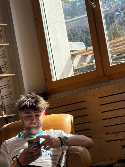 An indoor scene depicts a young male, approximately mid-20s, with dark, spiky hair and light facial stubble, seated in a tan leather armchair. He is smiling faintly, looking down at a light-colored smartphone held in both hands, featuring a clear case and a circular pop socket with a person's image. He wears a white short-sleeved t-shirt with light blue trim on the collar and "OM" branding on the chest, a gold chain around his neck, a watch with a translucent band on his left wrist, and multiple beaded and chain bracelets on his right wrist.

The subject is positioned with strong natural light entering from a large, multi-paned wooden-framed window dominating the background. To the left of the frame, a segment of a tall, narrow wooden shelf unit is visible. To the right, a wooden wall paneling with horizontal dark slits extends.

Through the window, an outdoor environment consistent with Briançon, France, is visible under bright daylight. A concrete or stone structure, possibly a bridge or elevated pathway, is discernible in the mid-distance above a cluster of evergreen trees. Below this, a wooden balcony or terrace structure with stacked timber planks is present. The setting suggests a sunny day in an alpine town.