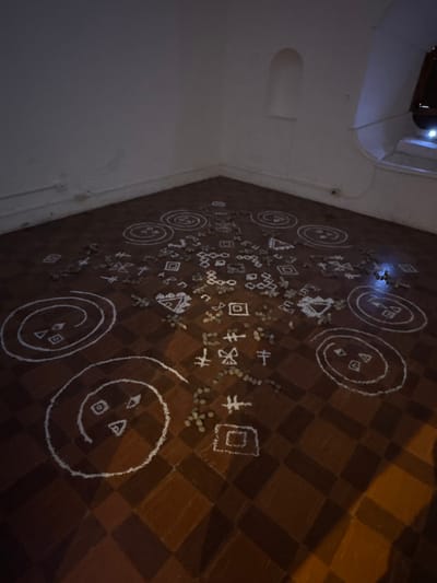 A floor displays geometric symbols drawn in white, including concentric circles, triangles, squares, and other abstract shapes. Small piles of round objects are scattered around the symbols. The floor has a checkered pattern, and the wall is light-colored with a small arched alcove. The lighting is dim, casting shadows across the room.
