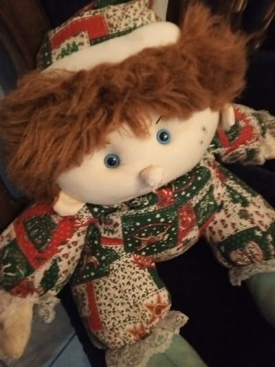 A soft, handmade doll with light blue eyes and brown hair. The doll's face is pale with a small, pointed nose. It wears a patchwork outfit in festive red, green, and white patterns. The outfit includes a matching hat with a white brim. Delicate lace trim adorns the edges of the sleeves and pants. The doll's body and limbs are plump and softly stuffed.