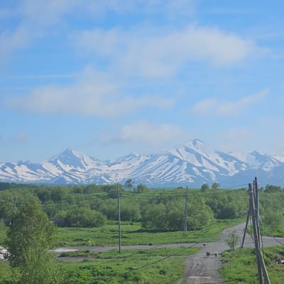 This vertical, eye-level media file shows a scenic mountain landscape under a blue sky with scattered clouds. The mountains are covered in patches of snow, suggesting a cooler climate or high altitude. In the foreground, a narrow, paved road winds through lush green vegetation. Telephone poles and power lines run parallel to the road, indicating a developed area. The vegetation appears to be a mix of trees and grass, with some wildflowers visible. The time of day appears to be daytime, judging by the bright sky and shadows. The weather seems to be clear and pleasant, with a gentle breeze suggested by the clouds. The overall mood is peaceful and serene, with a touch of rustic charm from the road and power lines. The location is identified as Sokoch, Russia, which aligns with the mountainous terrain and potential for snow-capped peaks. There is no visible text in the media file.