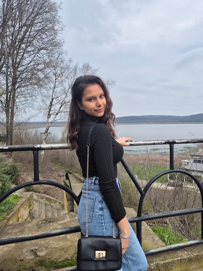A young woman is posing for a photo in what appears to be Srebarna, Bulgaria. She is leaning against a black metal railing, with a body of water and a landscape visible in the background. She is wearing a black turtleneck sweater, blue jeans, and a black purse with a gold chain and clasp. She has long brown hair and is wearing a bracelet on her left wrist. The railing shows signs of wear and age. Bare trees and vegetation are visible in the surrounding environment. The sky is overcast. Steps lead down from behind the railing. Buildings can be seen on the opposite shore in the background.