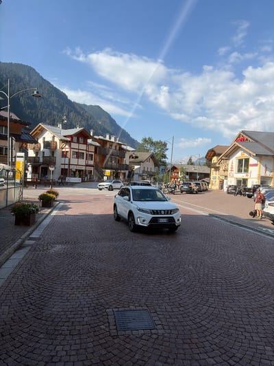 The image captures a street scene in Campitello di Fassa, Italy, under a bright, partly cloudy sky. The foreground is dominated by a cobblestone street with a curved pattern. A white Suzuki Vitara is parked in the middle of the street, facing towards the viewer. To the left of the Vitara, a row of buildings with traditional alpine architecture lines the street. These buildings feature wooden balconies, red and white facades, and grey roofs. Flower boxes with colorful blooms are visible in front of some of the buildings.
Further down the street, more buildings are visible, some with shops on the ground floor. A sign for "SPORT 365" is prominent on one of these buildings. Several other cars and motorcycles are parked along the street or in motion. A person with a dog is standing on the right side of the street near a parked car. In the background, a majestic mountain covered in pine trees rises above the town, with its peak partially obscured by clouds. The overall impression is a bustling, picturesque mountain town on a sunny day.