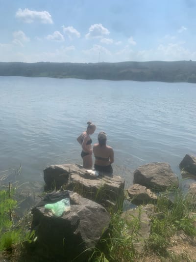 Two young women are wading in a body of water near the shore, which is lined with large rocks and vegetation. The woman closer to the shore is wearing a black bikini and has her hair tied up in a bun. She is looking away from the camera and appears to be interacting with the other woman. The second woman, partially submerged in the water, is also wearing a black bikini. She is facing the camera and has dark hair. The water is calm with ripples on its surface, reflecting the light blue sky and scattered white clouds. In the background, a distant, hazy shoreline with a gradual slope can be seen. The sun appears to be high in the sky, suggesting it is daytime, and the weather seems to be warm and pleasant. On the rocks near the shore, there are some discarded items, including a black piece of clothing and a bright green plastic bag, indicating a casual outing or recreation by the water. The overall scene depicts a relaxed summer day at a natural waterfront location.