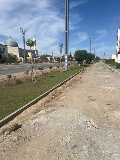 This image depicts a street scene in Balykchy, Kyrgyzstan. The photo is taken from an elevated perspective, looking down a wide, paved road that is bordered by a grassy median strip and a dirt path. The road appears to be under construction or repair, as the dirt path is rough and uneven, with loose gravel and debris.
Along the grassy median, young trees have been planted and are supported by wooden stakes. In the distance, buildings with white and light-colored facades are visible, one of which has a distinctive green dome, possibly indicating a religious building like a mosque. Several modern streetlights with decorative tops are also present along the road. The sky is a bright blue with scattered white clouds, suggesting a clear and sunny day. The overall impression is one of an urban environment undergoing development or renovation. There is no visible text in the image.