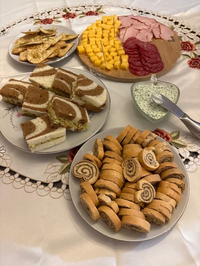 This scene, located in San Borja, Peru, displays a meticulously arranged spread of various appetizers and snacks on a light-colored table adorned with an embroidered tablecloth. The tablecloth features a repeating pattern of red roses, green leaves, and black and gold ornate borders. No individuals are present in the image.

From top-left, a round white plate contains numerous triangular flatbread chips or crackers, lightly seasoned with green specks, possibly dried herbs. To the right, a wooden cutting board presents an assortment of sliced meats and cheese: a pile of uniformly cubed yellow cheese with several toothpicks, a mound of thinly sliced pink deli meat (likely ham or turkey), and a stack of thinly sliced red cured sausage (salami or pepperoni).

Below the cutting board, a clear glass heart-shaped bowl holds a light green, creamy dip, interspersed with dark green flecks, suggesting a herb-based ingredient. A silver spreading knife rests partially submerged in the dip.

At the bottom right, a white plate is laden with numerous slices of a sweet rolled bread or pastry. Each slice reveals a distinct spiral pattern of light-colored dough and a darker, presumably sweet, filling.

To the left of the sweet rolls, another white plate holds several square-cut sandwiches. These sandwiches are notable for their two-toned bread, featuring a striped or marbled pattern of light and dark brown bread. The filling appears to be a light green, creamy salad-type spread, possibly egg salad with avocado or another herb-inclusive mixture.