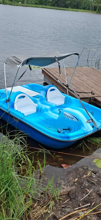 A bright blue pedal boat with white seats and a grey canopy is docked on a wooden pier next to a body of water. The boat has "Pelican" and "Voyage DLX" branding. Green reeds and aquatic plants are visible around the edges of the water and the boat. The background shows a treeline and a slightly overcast sky. The scene is located in Kohatu, Estonia.