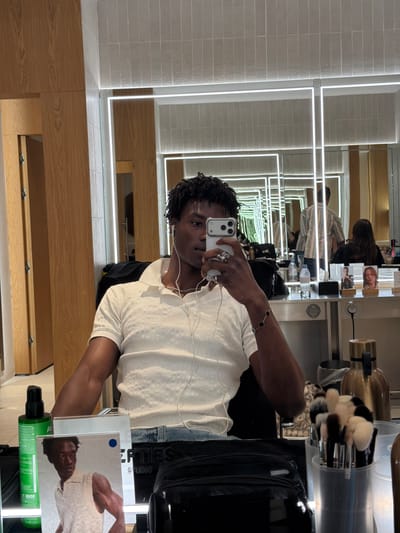 The image shows a young Black man taking a selfie in a brightly lit room, possibly a dressing room or salon, as indicated by the presence of makeup brushes, bottles of hair products, and mirrors. The room has wooden paneling and tiled walls, with several mirrors reflecting the scene. The man is wearing a white polo shirt and blue jeans, with white earphones in. He is holding up a smartphone to take the photo, wearing rings on his fingers and a bracelet on his wrist. In the background, other people are visible in the mirrors, and there are various objects on a counter, including bottles of water, hair products, and a gold-colored thermos. The overall impression is a casual, behind-the-scenes snapshot. There is no information within the image to suggest the location is Fibracolor, Spain.