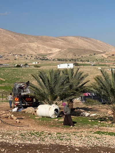 The image depicts an outdoor scene in the Arvot Hayarden Regional Council, Palestinian Territory, under a clear blue sky. In the foreground, four males are engaged with a dark-colored pickup truck. Two males are positioned on the truck bed, handling white, frame-like objects. One male stands beside the rear passenger side of the truck, wearing a grey hoodie. Another male is crouched or bent near the rear driver's side. The truck displays orange and yellow striped hazard markings on its tailgate.
To the right of this activity, a female figure stands under the shade of a date palm tree. She is dressed in a dark long skirt, a grey jacket, and a pink head covering, and is looking towards the left. A large, white, cylindrical tank lies horizontally on the ground near her, positioned in front of the palm tree. Behind the palm tree, a setup of solar panels is visible, with small items of clothing hanging to dry on a line nearby. Black hoses or pipes are present on the rocky, sparse ground in the immediate foreground.
The midground features patches of green vegetation, scattered small white modular structures, and other smaller buildings or tents. The background is dominated by a range of arid, light brown, undulating hills.