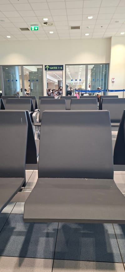 The image depicts an airport departure lounge in Rhodes, Greece. The scene is dominated by rows of dark grey, modular seating, with some chairs occupied by people waiting for their flights. Through a large glass window, a hallway with more seating and a view of a tarmac or other airport infrastructure can be seen. The lighting suggests it is daytime, with overhead lights illuminating the interior. A green "GATES 1-6" sign with an arrow pointing upwards is prominently displayed on the left side of the image. Another green exit sign with a person symbol is also visible on the wall. The overall atmosphere appears calm and orderly, typical of an airport waiting area. The people are varied in age and appear to be engaged in typical airport activities such as resting, talking, or looking at their phones. The tiling on the floor and the ceiling panels are modern and clean. The presence of the "Gates 1-6" sign strongly indicates this is an airport terminal.