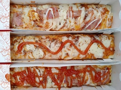 The image displays a close-up, top-down view of three rectangular flatbreads, resembling personal-sized pizzas, each housed in an individual white cardboard box. The boxes are stacked vertically, showcasing the toppings of each item.
The topmost flatbread is covered with a melted white cheese base, sprinkled with dried herbs, and features several slices of ham or similar processed meat. A white, creamy sauce, possibly mayonnaise, is drizzled in a wavy pattern over the toppings.
The middle flatbread also has a melted white cheese base and herbs. It is distinctly characterized by a single, undulating line of red sauce, likely ketchup, running across its length.
The bottom flatbread shares the melted cheese and herb base. Underneath a generous and more intricate crisscross drizzle of red sauce (ketchup), slices of meat, similar to those on the top flatbread, are visible.
The flatbreads appear freshly cooked, with slightly golden edges. The inner left wall of each white box is adorned with a decorative pattern of repeating red outlines of tomato clusters and leaves. The setting suggests food prepared for individual serving or takeaway. No specific text is visible, only the decorative graphic. The items are presented in a manner indicating they are ready for consumption, possibly in Kecamatan Panji, Indonesia.