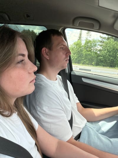 Two individuals, a woman and a man, are seated inside a car. The woman, in the foreground on the left, has light brown hair and is wearing a white t-shirt. She is looking to her left, away from the camera. The man, positioned to her right and slightly further back, is also wearing a white t-shirt and appears to be looking ahead. The interior of the car is visible, including the door panel and a grab handle on the ceiling. Through the car windows, a blurred background of green trees and a road can be seen, suggesting the car is in motion, possibly on a road in or near the city of Alnashi, Russia.