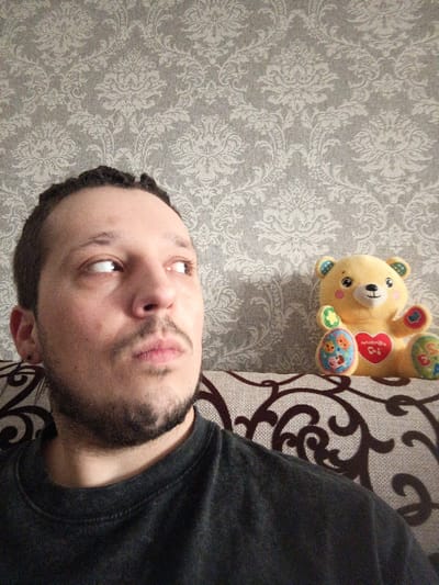 The image depicts an indoor scene in what is reported as Bessonovka, Russia. One adult male is positioned in the lower-left quadrant of the frame, with his head tilted slightly upwards and his eyes directed towards the upper-right, off-camera. He has dark, short curly hair, a beard, and a mustache, and wears a dark, possibly charcoal-colored, crew-neck t-shirt. A small black stud is visible in his left earlobe. To the right of the male, resting on a sofa, is a yellow plush teddy bear. The bear features colorful patches on its ears, hands, and feet, and a red heart-shaped patch on its chest containing text and musical notes. The background consists of a wall covered in light grey wallpaper with a repeating, light-toned damask pattern. A portion of the sofa, upholstered in a light fabric with dark brown, scrolling patterns, is visible in the lower right.