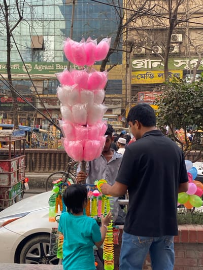 The scene depicts a street-level commercial interaction in an urban area, identifiable by Bengali script on signage, consistent with Narayanganj, Bangladesh. In the immediate foreground, a young child, viewed from the rear, wearing a turquoise t-shirt, stands facing a street vendor's display of colorful toys. To the child's right, an adult male, also viewed from the rear, wearing a black short-sleeved polo shirt and blue jeans, stands beside the child, observing or interacting with the toy selection.

The vendor, a male partially obscured by his wares, wears a grey patterned shirt and holds a vertical wooden pole. Attached to this pole are multiple individually plastic-wrapped servings of cotton candy, arranged in alternating tiers of pink and white, forming a prominent display resembling lotus flowers. Below the cotton candy, the vendor's display rack holds numerous brightly colored plastic toys, including spring slinkies in shades of yellow and green, and various bubble wands.

In the midground, the front section of a white vehicle, a car with a visible Toyota wheel hub, is parked or stopped on the left. A bicycle is discernible further back, behind the vendor. The background features a busy street with multi-story buildings. A prominent green sign for "Agrani Bank" is visible on a glass-fronted building. Additional commercial signage in Bengali script is present. Numerous overhead utility wires crisscross the upper portion of the frame. Bare tree branches are also visible. A cluster of multi-colored balloons (pink, green, orange, light blue) is partially visible on the far right, behind the adult male. Several pedestrians are visible further down the street, including one individual wearing a white skullcap.