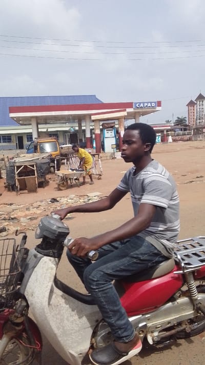 The image captures an outdoor street scene in Igbo Ukwu, Nigeria. In the immediate foreground, a young dark-skinned male, likely in his late teens or early twenties, is seated on a red and white motorcycle, facing right in a profile view. He wears a grey and white striped short-sleeve shirt, blue jeans, and black clogs with white soles and pink interior accents, with his hands positioned on the handlebars. The motorcycle has a wire basket attached to the front and a silver luggage rack over the rear fender.

The ground is an unpaved, reddish-brown dirt or sandy surface, littered with various debris including plastic and wood fragments. To the left, several makeshift wooden market stalls, some partially covered with black tarps, are visible. A yellow three-wheeled utility vehicle is parked behind these stalls. In the mid-ground, a person wearing a yellow garment is attending to a wheelbarrow filled with items.

Dominating the background is a "CAPAD" gas station, identifiable by its prominent sign, a blue roof, and a red and white canopied structure supported by multiple pillars, under which fuel pumps are present. Other buildings with light blue exteriors are visible behind the gas station. To the far right, a tall building with distinctive multiple towers or spires, consistent with a church, stands in the distance. The sky is overcast or hazy grey, with overhead power lines visible across the upper portion of the frame.