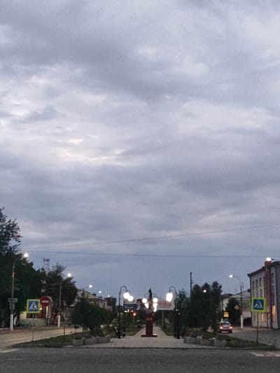 The image depicts a street scene in Slavgorod, Russia, during twilight. The sky is overcast with thick clouds. In the foreground, a paved road is visible, with a sidewalk to the left and right. Several streetlights with illuminated bulbs line the sidewalk. A pedestrian crossing sign is present on the left side of the road. Trees and buildings are visible in the background, some with illuminated windows. A monument is centrally located on the right side of the sidewalk, and a car is driving on the road in the mid-ground. The overall atmosphere is subdued due to the low light conditions.