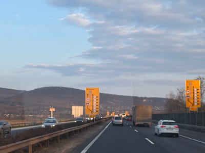 The image depicts an active multi-lane section of the M1 motorway, located near Tatabánya, Hungary, with traffic proceeding eastbound towards Budapest. The viewpoint is from within a vehicle traveling in one of the left lanes. The road surface is asphalt with distinct white lane markings. Vehicles observed in motion include a white station wagon in the rightmost lane, a grey semi-trailer truck in the adjacent middle lane, and multiple passenger cars distributed across the lanes, including a silver sedan in the opposing direction beyond the median. Two prominent yellow rectangular road signs are positioned on the right shoulder; both display "Budapest" at the top, illustrate upcoming lane configurations with multiple arrows, and include a "100 m" distance marker at the bottom. A dark grey sound barrier runs parallel to the motorway on the right, and a metal guardrail delineates the median strip. The background features a range of dark, tree-covered hills under a partly cloudy sky, with a telecommunication tower visible on the summit of the leftmost prominent hill. Low vegetation and bare ground are present in the median and along the road shoulders. No pedestrians are visible.