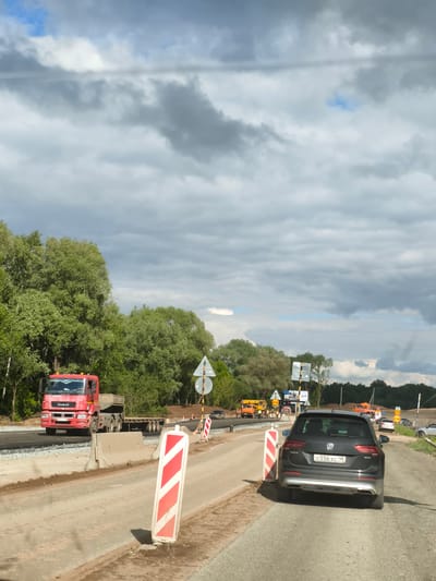 The image depicts a road construction site in Kabanikha, Russia, under a cloudy sky with glimpses of blue. The foreground features a temporary dirt road section flanked by red and white striped warning markers and concrete barriers.

On the left, a large red KAMAZ truck with a flatbed trailer is driving on a newly laid asphalt section of the road. The truck has "KAMAZ" branding visible on its cab and grille, and a Russian license plate. A person is visible in the driver's seat. Adjacent to the road, dense green trees line the landscape.

In the mid-ground, beyond the barriers, construction activity is evident with yellow heavy machinery and several workers in orange safety vests. Various road signs are scattered, including one indicating a "30" speed limit and others with Russian text, such as "ПЕРЕЕЗД" (crossing) and "ЛЮМ".

In the right foreground, a dark grey Volkswagen Tiguan SUV is parked or moving slowly, facing away from the viewer. The vehicle displays "Volkswagen Tiguan" badging and a clear Russian license plate "o 556 xc 18". The sky above is largely covered by grey clouds, though some patches of clear blue sky are visible. The overall scene indicates daytime with ongoing road work.