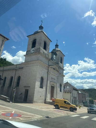 The image shows a view of the Église Saint-Pierre in Morbier, France, on a sunny day. The church is made of stone and features two bell towers with clocks and crosses on top. The facade has arched windows and the Latin inscription "QUIS UT DEUS" above a door, along with the year "1836". A yellow postal van is parked on the street in front of the church, and a dark-colored car is also visible on the road. The street has pedestrian crosswalk markings and a "no parking" sign. In the background, there are residential buildings and a tree-covered hill under a blue sky with scattered clouds. The overall scene depicts a typical village square in France.
