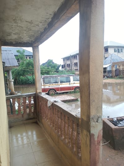 The image depicts a flooded street scene in Ihiala, Nigeria. A white and red bus, partially submerged in brown water, is visible in the middle ground. Buildings with pitched roofs are situated in the background, with some appearing to be multi-story structures. The foreground shows a tiled balcony or patio with a decorative railing. A concrete pillar supports an overhead structure. The water level suggests a significant flood event, with debris visible in the water. The sky is overcast.