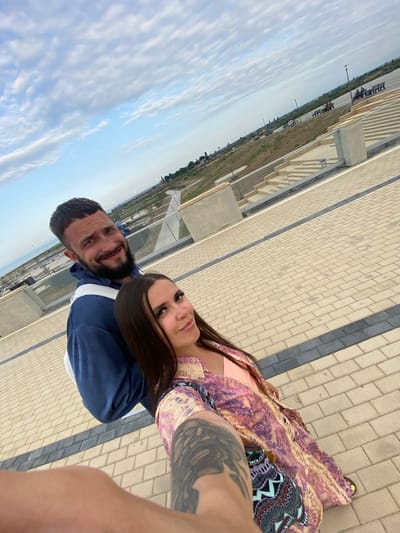 A man and a woman are taking a selfie outdoors, with the city of Arablyar, Russia visible in the background. The man, with a beard and a short haircut, is wearing a blue hoodie. The woman, with long dark hair, is wearing a pink top and a colorful patterned dress. They are standing on a paved area with a tiled walkway and stairs. The sky is partly cloudy. The location appears to be a public space or park, possibly with a view of the sea, given the distant coastline and the open expanse.