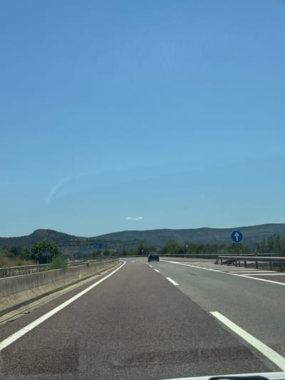 The image captures a daytime scene on a highway, likely in the vicinity of Canals, Spain. The perspective is from inside a vehicle, looking forward. The highway has multiple lanes separated by white lines. The road surface appears to be asphalt with some reddish-brown aggregate. On the left side of the road, a concrete barrier separates the highway from an embankment with some vegetation. On the right side, a metal guardrail is visible. The sky is a clear, bright blue with a faint wispy cloud formation. In the distance, rolling hills covered in trees and vegetation form the horizon. There are a few road signs visible, including one with an upward-pointing blue arrow and another triangular sign with a blue circle and arrow. A single dark-colored car is traveling ahead on the highway. The overall environment suggests a rural or semi-rural highway setting under fair weather conditions.