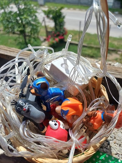 A close-up, high-angle shot captures a small, light-brown woven basket filled with an assortment of tangled clear string lights and several small, colorful plastic figurines, set outdoors in Bregovo, Bulgaria.
The basket holds approximately six distinct toy figures. Among them are characters resembling the Paw Patrol pups, including one in a blue uniform with a police hat (Chase), another in an orange uniform with a construction hat (Rubble), and one in a red uniform with a firefighter hat (Marshall). Additionally, a black and grey figure with a red nose, resembling "The Little Mole" character, is visible, holding a small shovel. These figurines are nestled within and partially obscured by a substantial tangle of transparent-wired LED string lights. A small, white rectangular plastic component, likely a battery pack or control unit for the lights, is also visible amidst the wires.
The basket rests on a textured, weathered wooden surface, possibly a railing or plank. The background is softly blurred, revealing lush green foliage, some vibrant red flowers, and a glimpse of a paved road or path further in the distance. The scene is illuminated by bright natural sunlight, creating distinct shadows and suggesting a clear, sunny day. The overall impression is one of items stored or arranged casually in an outdoor setting. No legible text is visible on any of the objects.
