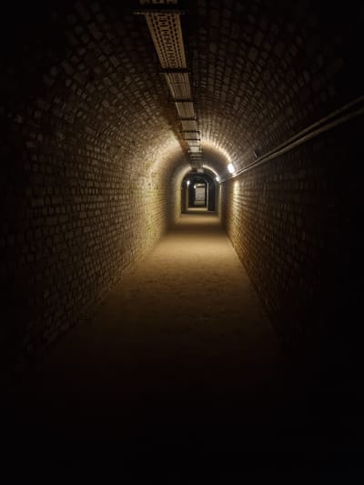 The image shows a dimly lit tunnel, likely a former water tunnel in Homburg, Germany. The tunnel walls and floor are constructed of brick or stone, and a metal railing runs along the left side. Lights are spaced intermittently along the tunnel's ceiling, casting a faint glow on the surrounding surfaces. The tunnel narrows towards the vanishing point, eventually becoming a rectangular opening. To the right, the tunnel wall is smoother, possibly concrete, and fades into darkness. No people are visible.