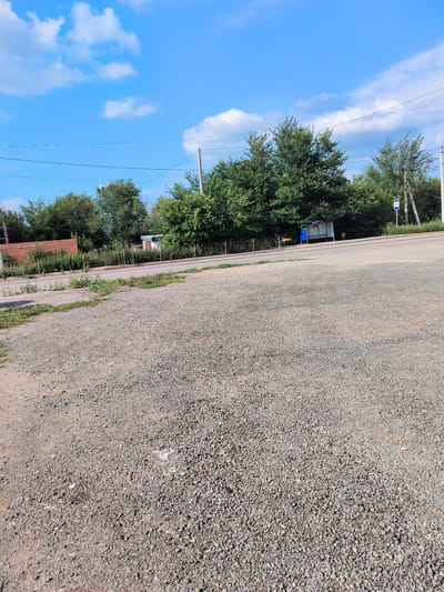 The image captures a sunny day in Pokhvistnevo, Russia. The foreground is dominated by a gravel surface, appearing to be a roadside or parking area. Beyond this, a paved road runs horizontally across the mid-ground. On the opposite side of the road, a line of mature green trees forms a dense backdrop. Visible behind the trees are some structures, including a brick building on the far left and a bus stop shelter with a blue roof and side panels further to the right, identifiable by a blue bus stop sign. Power lines are strung across the sky, supported by utility poles, one of which is visible on the right side of the frame. The sky is a bright blue with scattered white clouds. The overall scene depicts an urban or semi-urban environment, possibly near a main thoroughfare or residential area.