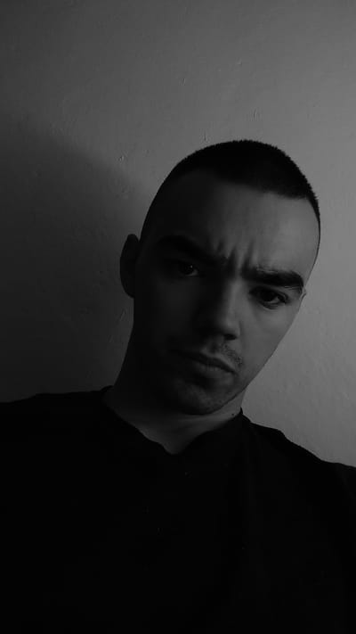 A low-angle, black and white, close-up photograph shows the upper body and head of a young adult male against a textured, off-white wall. The man has a shaved head and is wearing a dark t-shirt. His face is partially illuminated by a light source coming from the right, casting strong shadows. His eyebrows are furrowed, and his expression appears serious or intense. The lighting emphasizes the contours of his face and the texture of the wall. The image was captured in Sosnowiec, Poland.