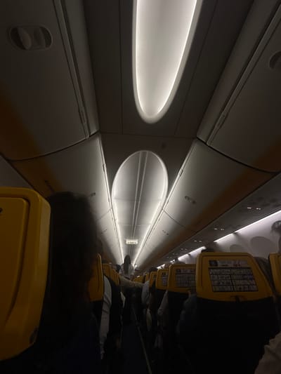 The image depicts the interior of a Ryanair airplane. Passengers are seated in yellow seats along the aisle. Overhead compartments line the ceiling of the aircraft, featuring illuminated recessed lighting. An "EXIT" sign is visible. The location is implied to be in Luqa, Malta, based on Ryanair's operation from Malta International Airport located in Luqa.