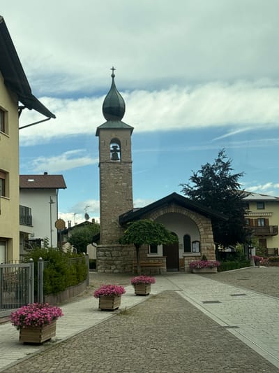 The image shows a view of the village of Fai della Paganella, Italy, on a cloudy day. In the foreground, a cobblestone street is lined with flower planters filled with pink petunias. To the left, a yellow building and a fence are visible. To the right, a small stone church with a bell tower stands prominently. The bell tower is made of stone with a dark green, pointed roof topped with a cross. A bell is visible inside the tower's opening. A tree with dark foliage stands behind the church. Further in the background, several other buildings, including a yellow building with balconies and solar panels, are visible. The sky is partially cloudy with some patches of blue sky showing through.