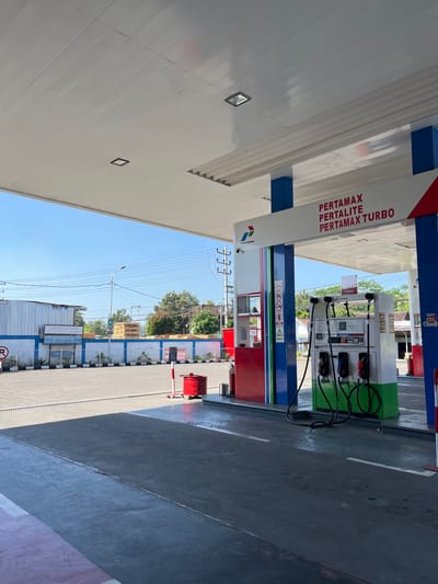 The image depicts a Pertamina gas station in Purworejo, Indonesia. The scene is captured from a low angle, looking up towards the canopy of the gas station. Under the canopy, a Pertamina gas pump is visible, with various fuel options like Pertamax, Pertalite, and Pertamax Turbo advertised. The pump has several nozzles ready for use. To the left of the pump, there is a red fire extinguisher. In the background, there is a paved area with a clear blue sky overhead. A white building with a blue stripe along the top is visible behind a low blue and white wall. There are also trees and power lines in the distance, suggesting an urban or suburban environment. The overall impression is one of a clean and functional service station.