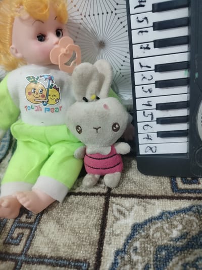 The image shows a doll with blonde hair and a pacifier in its mouth, wearing a white shirt with an orange cartoon character print and green pants. A small stuffed bunny wearing a pink and black striped dress is next to the doll. To the right of the doll and bunny is a small toy keyboard with numbers written on the keys. The items are placed on a rug with a patterned design. The background consists of a white surface with a radiating pattern. The image appears to be a close-up shot taken indoors. There is no indication of the city of Cholpon, Kyrgyzstan in the image.