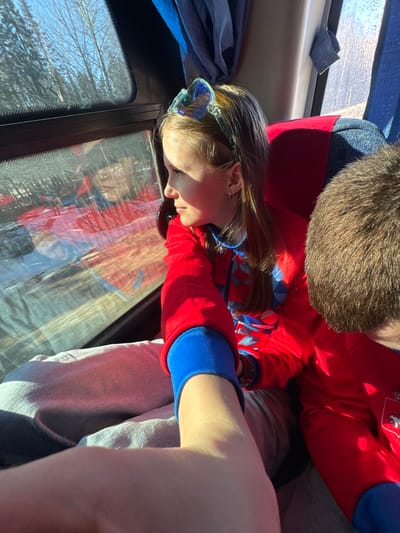 A young girl with light brown hair and a blue headband is looking out the window of a vehicle. She is wearing a red and blue outfit. A person with short brown hair is beside her, also wearing a red and blue outfit. The window shows reflections of the people in the car and trees. The exact location in the city of Челва, Russia, cannot be determined from the image.