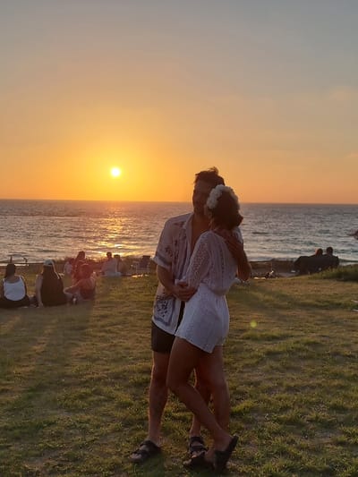 A couple is embracing on a grassy area overlooking the Mediterranean Sea at sunset in Palmachim, Israel. The sun is setting on the horizon, casting an orange glow across the sky and water. Several people are scattered on the grass in the background, appearing to be watching the sunset. The couple in the foreground is close together, with the man's arms around the woman. The woman is wearing a white lace dress and a flower crown, suggesting a celebratory occasion. The scene conveys a romantic and peaceful atmosphere.