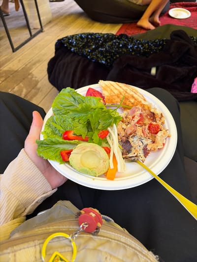 A person's hand, wearing a beige corduroy shirt, holds a white paper plate filled with food. The plate contains a salad with lettuce, red bell pepper, and parsley, half an avocado, some shredded white vegetables and orange carrot sticks. To the right of the salad is a portion of scrambled eggs with tomatoes and bacon, and a grilled sandwich cut in half. A yellow fork is also visible on the plate. The person is sitting on a black surface. In the background, part of a wooden floor and a black fabric with sequins are visible. The scene does not provide any specific details that would allow for identification of the city of Shebanovo, Russia.