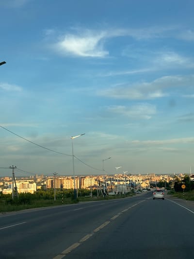 A high-angle shot captures a paved road leading into the city of Plodopitomnik, Russia, under a partly cloudy sky. The road, marked with yellow and white lines, curves gently to the right as it approaches a sprawling urban landscape. On either side of the road, grassy verges are dotted with trees and telephone poles. In the distance, a cityscape unfolds, characterized by numerous apartment buildings and other structures, some of which are illuminated by the golden light of what appears to be sunset. A silver car is visible on the road ahead, traveling in the same direction as the viewer. A speed limit sign, indicating 40, is posted on the right side of the road. The scene conveys a sense of approaching dusk in a residential area.