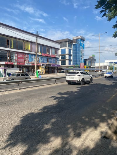 The image depicts a street scene in Parkent, Uzbekistan, on a sunny day with a blue sky and scattered clouds. In the foreground, a grey station wagon is parked alongside a road, and a modern white SUV is driving away from the camera. The road itself appears to be asphalt with some wear and tear, and is divided by a metal guardrail.
On the left side of the street, there is a multi-story commercial building with large windows. Signs on the building indicate various businesses, including "Optom Market," "KONG ZIYO OQUV MARKAZI," and "AVTO DOKTOR." A blue and white vertical sign is visible on a taller building further down the street, with the text "BURKON OTA." Several other smaller commercial buildings and structures are visible in the background.
Trees with green leaves are present on the right side of the image, casting shadows on the road and sidewalk. The environment appears to be an urban commercial area. There are no clearly visible people engaged in significant actions or interactions, though some individuals may be present within the buildings or on the sidewalk. The overall impression is a typical day in a commercial district of a Uzbek city.