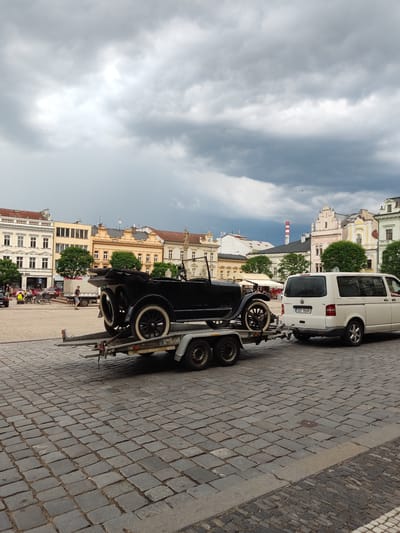 In Kolín, Czechia, a black vintage car is being transported on a trailer towed by a white Volkswagen Transporter van. The scene unfolds on a cobblestone square in front of a row of historic European buildings. The sky is filled with dramatic, heavy gray clouds, suggesting an impending storm. Several people are visible in the background, some seated at outdoor tables, others walking. The vintage car, with its large white-rimmed wheels and open-top design, is the primary focus, positioned prominently on the trailer. The van is a modern vehicle, creating a contrast between old and new. The architecture of the surrounding buildings features classic European styles with arched windows and ornate facades, indicative of a historical city center.